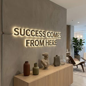 Op de afbeelding: Een wandbord met verlichte 3D-letters die SUCCESS COMES FROM HERE spellen in een warme gloed. Daaronder staat een lichthouten kast met een terracotta vaas, een saliegroene vaas, een klassieke buste, een grijze vaas en een doorzichtige glazen vaas.