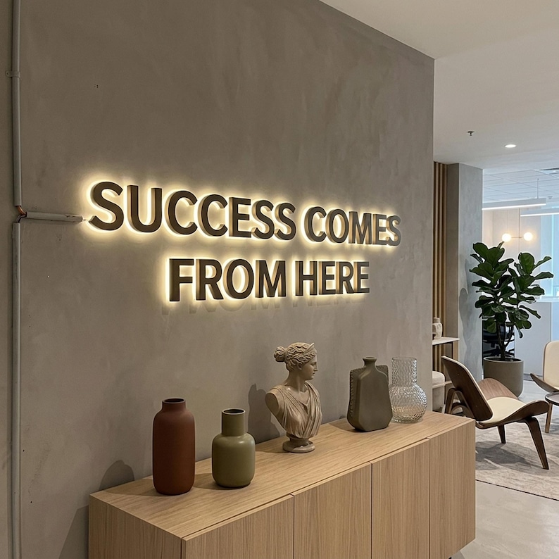Op de afbeelding: Een wandbord met verlichte 3D-letters die SUCCESS COMES FROM HERE spellen in een warme gloed. Daaronder staat een lichthouten kast met een terracotta vaas, een saliegroene vaas, een klassieke buste, een grijze vaas en een doorzichtige glazen vaas.