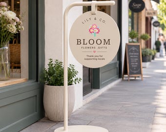 Op de afbeelding: Een beige rond metalen bord dat aan een bijpassende standaard hangt. Het bord bevat de tekst LILY & CO. BLOOM FLOWERS .GIFTS, een kleurrijke bloemafbeelding, een klein hartje en de tekst Thank you for supporting locals in donkere letters.