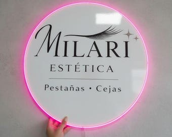 以下が含まれることがあります： ピンクのネオンの縁取りがある円形の白い看板。黒い文字で「Milari Estética」「Pestañas • Cejas」と書かれ、まつげのグラフィックと2つの小さな四芒星のアイコンがデザインされています。ブランド名がスタイリッシュなフォントで表示されています。
