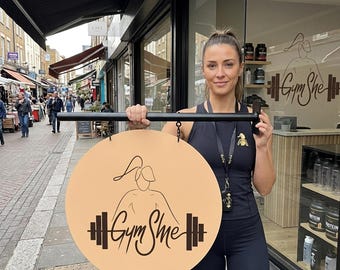 Op de afbeelding: Een rond, lichtbruin hangend bord met een zwarte lijntekening van een persoon met een paardenstaart en een halter, met de tekst Gym She in een gestileerd lettertype, opgehangen aan een zwarte horizontale metalen stang.