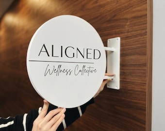 以下が含まれることがあります： 白い円形の壁掛け看板。太字のサンセリフ体で黒く「ALIGNED」と書かれ、その下に水平線があり、さらにその下に黒い筆記体で「Wellness Collective」と記されている。白いブラケットで固定されている。
