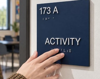 以下が含まれることがあります： 173 AおよびACTIVITYという白い文字が記された正方形の濃い青色の壁掛けサイン。各行のテキストの下には、触知可能な隆起した点字が配置されています。サインは明るい色の壁面に取り付けられています。