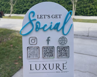 以下が含まれることがあります： 青い筆記体で「LET'S GET Social」と書かれたアーチ型の白い看板。3つの銀色のソーシャルメディアアイコンと3つのQRコードが配置されています。下部には黒い文字で「LUXURÉ REAL ESTATE」と記され、黒い台座の上に設置されています。