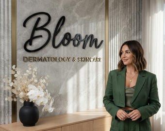 以下が含まれることがあります： グレーの大理石の壁面に、黒い筆記体の「Bloom」と金色の「DERMATOLOGY & SKINCARE」の文字が掲示されています。白い花を生けた黒い花瓶が明るい木製のキャビネットの上に置かれ、その横にダークグリーンのブレザーとパンツを着用した人物が立っています。