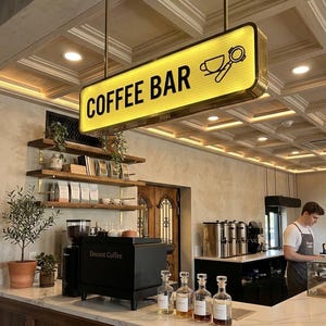 Może przedstawiać: Wnętrze kawiarni z podświetlanym szyldem z napisem "COFFEE BAR". Znak zawiera filiżankę kawy i ikonę portafiltra. Bar posiada czarny ekspres do kawy, szklane butelki i baristę.