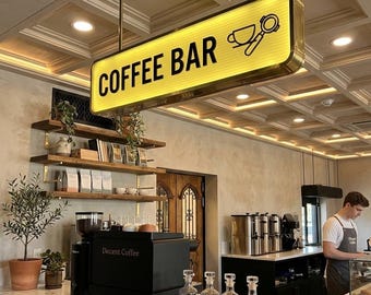 以下が含まれることがあります： 「COFFEE BAR」と書かれた照明付きの看板があるコーヒーバーのインテリア。看板にはコーヒーカップとポルタフィルターのアイコンがあります。バーには黒いエスプレッソマシン、ガラス瓶、バリスタがいます。