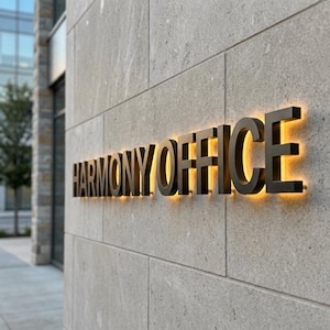 Op de afbeelding: Een driedimensionaal wandbord met de zwarte tekst HARMONY OFFICE gemonteerd op een lichtgrijze stenen muur. De letters zijn verlicht met een warme gele gloed, wat een duidelijk silhouet cre&euml;ert tegen het gestructureerde oppervlak.