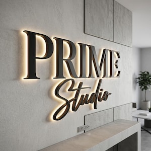 Peut inclure: Une enseigne murale avec le texte noir PRIME Studio rétroéclairé créant un effet de halo lumineux. Sous le texte principal, une petite plaque rectangulaire affiche le texte EST. 2024 dans une police sans empattement épurée.