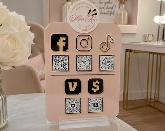Op de afbeelding: Een lichtroze acryl bord voor sociale media met een rond logo bovenaan met de tekst Beauty By Ollvia Aose Est. 2021. Het bord toont gouden en zwarte iconen voor Facebook, Instagram, TikTok, Venmo en Cash App, vergezeld van bijbehorende QR-codes.