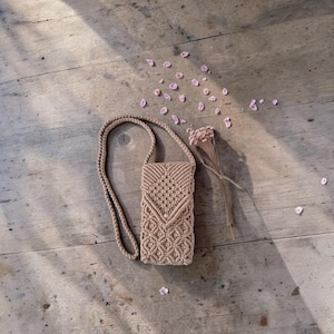 Puede incluir: Un bolso bandolera de macramé beige para teléfono con una correa larga. El bolso presenta un patrón geométrico en la parte delantera y un cierre de solapa. El bolso está hecho de material tejido y se muestra sobre una superficie de madera con pétalos de flores rosas y una flor seca.