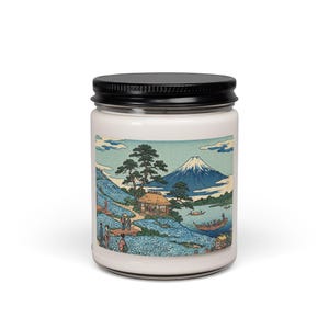 Op de afbeelding: Een witte kaarsenpot met een zwarte deksel, met een Japans landschapsontwerp. De afbeelding toont een berg, bomen, een kleine hut en mensen bij water. Het ontwerp is in tinten blauw, groen en bruin.