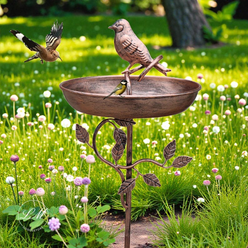 May include: A bronze-colored bird bath with a decorative leaf design. The bird bath has a bowl shape and is supported by a slender, ornate stand. Several birds are perched on the bird bath and flying nearby, set against a backdrop of green grass and flowers.