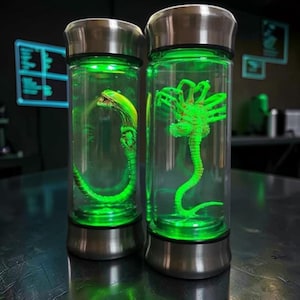 Op de afbeelding: Twee glazen bekers met roestvrijstalen deksels en bodems, elk met een neon groen alien figuur. De figuren zweven in een heldere vloeistof en de bekers worden verlicht met groene LED-lampjes.