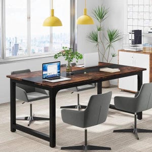 May include: A large, rectangular wooden desk with a black metal frame, paired with four grey swivel chairs. Two laptops are open on the desk. The room has large windows, yellow pendant lights, and a white cabinet.