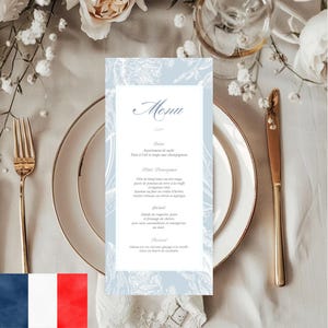May include: A formal table setting with a menu card. The menu card has a light blue background with white floral illustrations and the word "Menu" in elegant script. A gold fork and knife are placed on either side of the white plate.