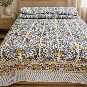 Könnte beinhalten: Eine gesteppte Tagesdecke mit einem sich wiederholenden Blumenmuster in Schieferblau, Senfgelb und Anthrazit auf weißem Grund. Das Design umfasst eine breite dekorative Bordüre mit einem wiederkehrenden geometrischen und floralen Motiv.