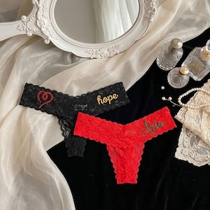 May include: Black and red lace thong underwear. The black thong has the word "hope" embroidered in gold and a red heart design. The red thong has the name "Aria" embroidered in gold. The underwear is displayed on a black surface.