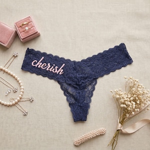 May include: Navy blue lace thong underwear with the word "cherish" embroidered in pink. The underwear is surrounded by wedding accessories, including a ring box, jewelry, and a bouquet of white flowers.