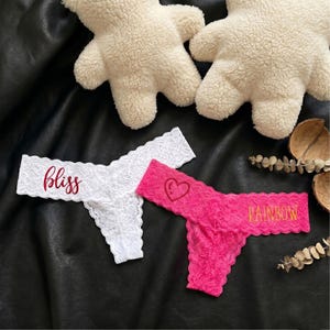 May include: White and pink lace thong underwear. The white thong has the word "bliss" in red script. The pink thong has a heart and the word "rainbow" in gold script. Two plush bear-shaped pillows are in the background.