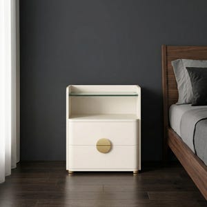 May include: Cream-colored bedside table with two drawers and a glass shelf. The drawers have a gold-colored, half-circle handle. The table has gold-colored feet and is next to a wooden bed frame.
