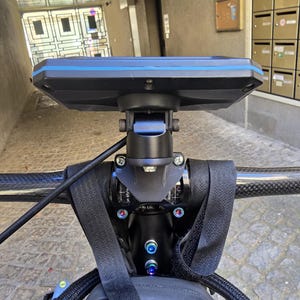 May include: A black and blue bicycle computer mounted on the handlebars. The computer has a rectangular screen with a blue border. The handlebars are black and have a carbon fiber appearance. The mount is secured with black straps.