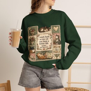 Op de afbeelding: Een donkergroen sweatshirt met lange mouwen en een patchwork-stijl afbeelding met diverse katten, bloemmotieven en de tekst: I work hard so my cat can have a better life (spoiler: they nap a lot) en CEO of Naps & Snacks.