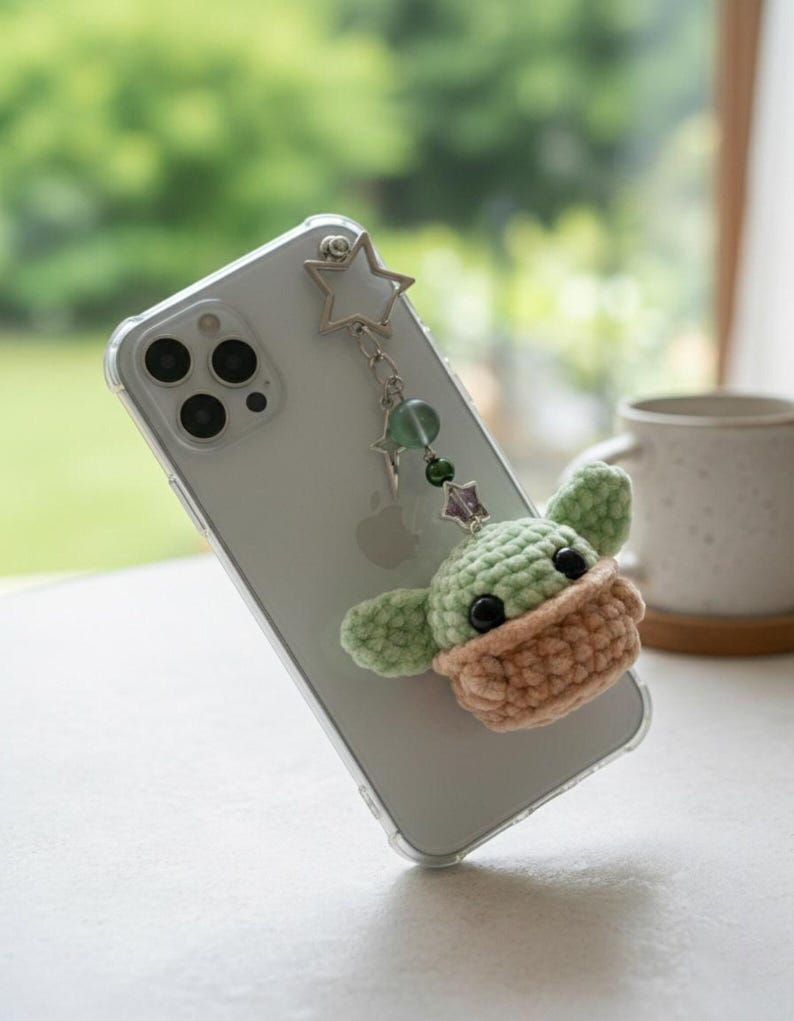 Op de afbeelding: Een doorzichtige telefoonhoes met een gehaakte Baby Yoda-bedel. De bedel is groen en bruin, met zwarte ogen. Een zilveren ster- en kralenketting verbindt de bedel met de telefoon. De telefoon ligt op een wit oppervlak.