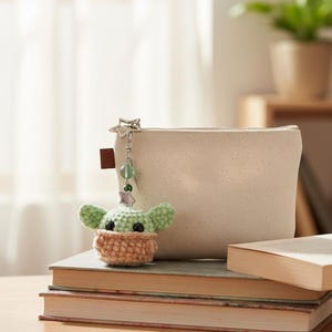Op de afbeelding: Een gehaakte Baby Yoda sleutelhanger met een lichtgroen hoofd en een bruin lichaam hangt naast een beige etui met ritssluiting. De sleutelhanger heeft een zilveren sterbedel en groene kraalaccenten. De items liggen op een stapel boeken.