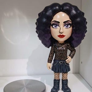 May include: A stylised figurine with voluminous dark purple hair, light skin, and purple eyes. The figure wears a brown leopard print long-sleeve top, a black pleated skirt with a heart-shaped belt buckle and chain detail, fishnet stockings, and black boots.