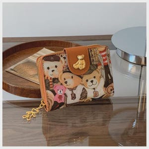 Pode incluir: Uma pequena carteira com zíper com um padrão repetido de ursinhos de pelúcia em várias roupas. A carteira tem uma aba de couro marrom com um emblema de urso dourado e um puxador de zíper dourado. O esquema de cores geral é bege, marrom e bronzeado.