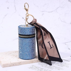 May include: A blue cylindrical keychain case encrusted with rhinestones, featuring a black zipper and a gold-tone keyring. A tan and black patterned scarf is attached to the keyring. The case is resting on a light-colored surface.