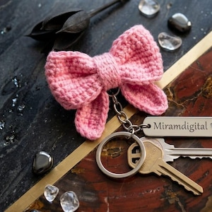 Puede incluir: Un llavero de lazo rosa tejido a ganchillo con una etiqueta plateada que dice "Miramdigital" y dos llaves. El llavero está sobre una superficie decorativa con un patrón de piedra negra y marrón.