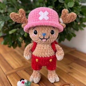 May include: A handmade crochet plush toy of a cartoon character, wearing a pink hat with a white cross, red shorts, and a small backpack. The plush has brown antlers and a blue nose. A small bracelet and miniature accessories are also present.