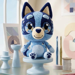 Peut inclure: Une peluche crochetée représentant un chien bleu avec de grands yeux brodés, des oreilles pointues et une tache ventrale bleu clair. La figurine présente des textures de fil bleu foncé et bleu clair, un petit nez foncé et un sourire cousu, posée sur un socle bleu clair.