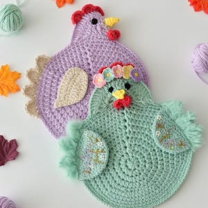 May include: Two handmade crochet chicken-shaped trivets. One is lavender with a red comb and yellow beak. The other is mint green with a floral crown and embroidered details. Autumn leaves and yarn balls surround the trivets.