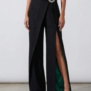 May include: Black wrap-style trousers with a high waist and a decorative pearl and rhinestone buckle. The trousers feature a slit on each leg, revealing a green lining and a decorative hem.