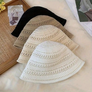May include: Four open-weave bucket hats in black, tan, and off-white. The hats are made of a knitted material with a wide brim and a slightly rounded crown. The hats are stacked on top of each other, showcasing the different colors and textures.