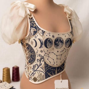 Peut inclure: Un corset crème avec un motif céleste représentant des lunes, des constellations et un aigle. Le corset a un devant lacé, des manches blanches transparentes et est orné de nœuds et de perles. L'étiquette indique "MairamDigital DIGITAL PATTERNS & DESIGNS."