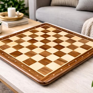 May include: A square wooden chess board with a light and dark brown checkered pattern. The board has a dark brown border with inlaid markings. The board is displayed on a light-colored table.