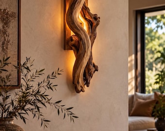 Op de afbeelding: Een wandlamp bestaande uit een verticaal geplaatst stuk natuurlijk, gedraaid drijfhout. Het hout is gemonteerd op een rechthoekige basis en wordt van achteren verlicht, wat een warme gloed op de muur werpt.
