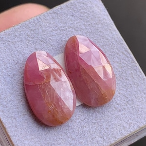 Puede incluir: Dos gemas rosas facetadas, de forma ovalada. Las gemas se exhiben sobre una superficie gris claro, mostrando sus superficies pulidas y patrones internos. Las piedras tienen un tono rosa rojizo con áreas más claras y oscuras, sugiriendo variaciones naturales.