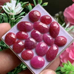 Puede incluir: Una colección de rubíes de color rojo rosado, con formas ovaladas y redondas, dispuestos sobre una base cuadrada de color violeta claro. Cada piedra preciosa presenta una superficie pulida y multifacetada que refleja la luz.