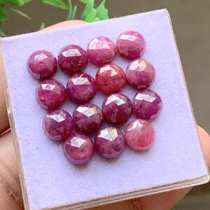 Pode incluir: Uma coleção de catorze pedras preciosas redondas, facetadas e de um vermelho rosado profundo, dispostas em grade sobre uma almofada de exibição quadrada de cor violeta claro. Cada pedra apresenta um corte geométrico simétrico.