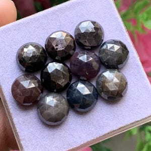 Puede incluir: Una colección de nueve piedras preciosas redondas y facetadas en tonos gris oscuro, negro y morado intenso. Cada piedra presenta un patrón geométrico de talla rosa con múltiples facetas triangulares, dispuestas sobre una base cuadrada de color lila claro.