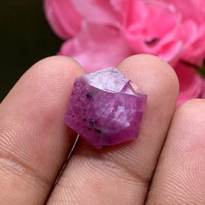 Könnte beinhalten: Ein sechseckiger, facettierter Edelstein in einem tiefen Magenta- und Rosaton mit sichtbaren inneren Einschlüssen und dunklen Sprenkeln im gesamten Stein. Der Edelstein zeigt eine polierte Oberfläche mit scharfen Kanten und Struktur.