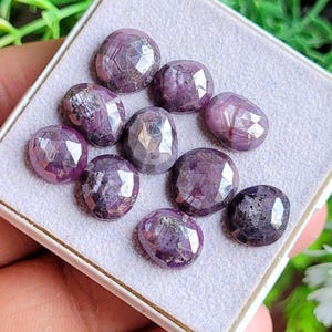 Puede incluir: Un conjunto de gemas facetadas, de forma ovalada, en tonos púrpura y rosa. Las piedras, probablemente rubíes, se exhiben en una bandeja blanca, mostrando sus superficies pulidas y patrones internos. Ideales para joyería.