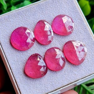 May include: Six faceted, oval-shaped pink gemstones arranged in a display box. Each stone features a polished surface with geometric cuts that reflect light, showcasing a vibrant, saturated magenta hue across all individual pieces.