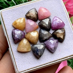 Puede incluir: Una colección de doce piedras preciosas triangulares facetadas en varios colores, incluyendo rosa, amarillo, morado, gris oscuro y marrón. Las piedras están dispuestas en un grupo sobre una superficie texturizada gris claro en una caja cuadrada.