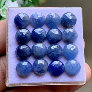 Peut inclure: Un ensemble de seize pierres précieuses saphir bleu, rondes et facettées, disposées en une grille de quatre par quatre sur un présentoir carré de couleur violet clair. Chaque pierre présente une taille brillant avec des facettes.